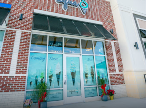 Storefront of an ice cream shop with large windows, "Coming Soon" signs, and decorative plants at the entrance.