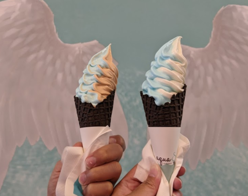 Two hands hold ice cream cones with swirled blue and brown soft serve, set against a backdrop of angel wings.