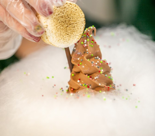 A hand adds a toasted marshmallow to a chocolate soft serve ice cream on a bed of cotton candy, sprinkled with colorful dots.