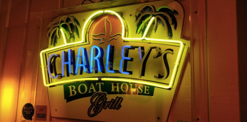 Neon sign for Charley's Boat House Grill, featuring palm trees and vibrant colors against a wooden background.