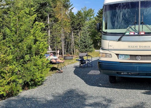 A person relaxes in a chair beside a parked RV in a wooded campsite.