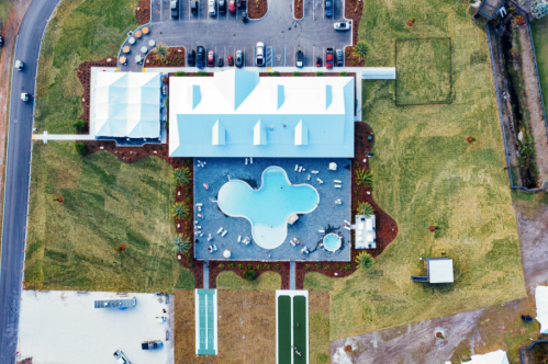 Aerial view of a hotel with a pool, surrounded by green grass and parking, near a road.