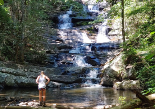 A person stands by a serene waterfall surrounded by lush greenery and rocky terrain.