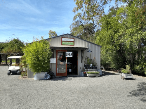 A small metal building with a sign reading "Visitor Center" and an "Open" sign, surrounded by greenery and gravel.