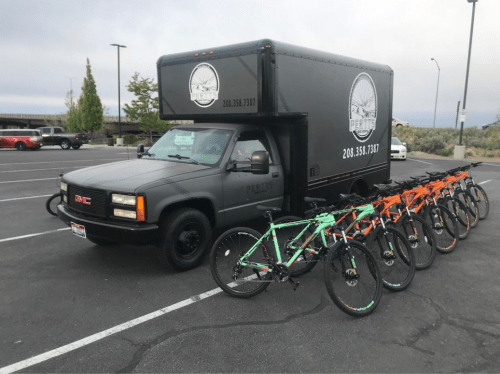 A black truck parked in a lot, with several bicycles lined up in front of it.