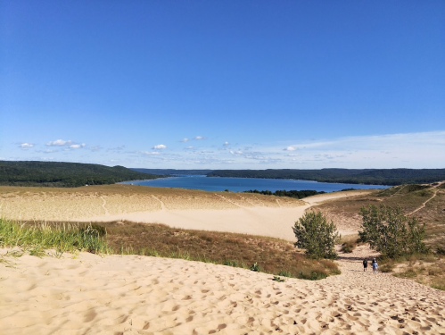 A sandy dune landscape with a lake in the background and green hills under a clear blue sky.