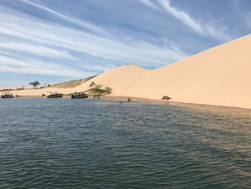 A serene landscape featuring a sandy dune beside a calm body of water, with people enjoying the beach and boats nearby.