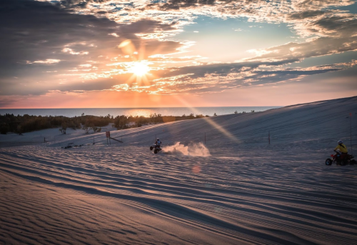 Two ATVs ride across sandy dunes at sunset, with the sun casting rays through scattered clouds over the ocean.