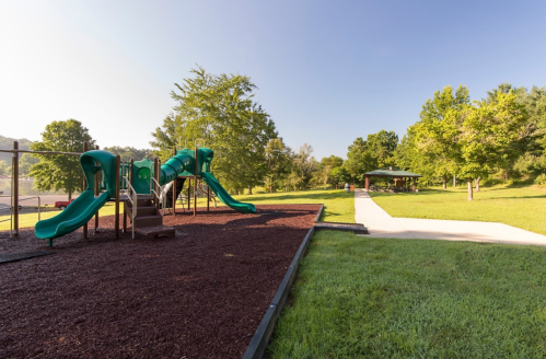 A playground with green slides and a nearby picnic area, surrounded by trees and a clear blue sky.