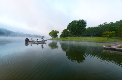 A fisherman stands on a boat in a misty lake, surrounded by lush greenery and calm waters.