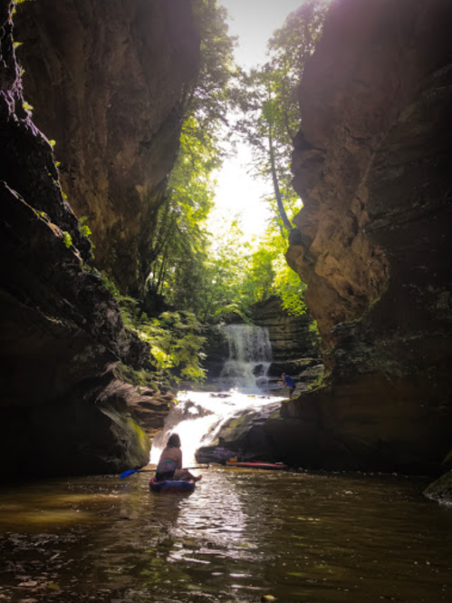 A serene scene of two people kayaking in a narrow canyon with a waterfall and lush greenery surrounding them.