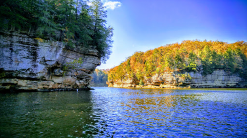 Scenic view of a calm river surrounded by rocky cliffs and autumn-colored trees under a clear blue sky.