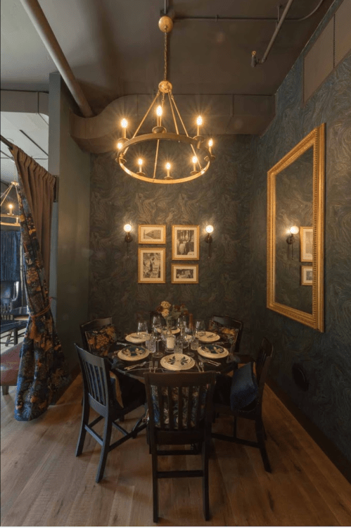 A cozy dining area with a chandelier, dark floral wallpaper, and a table set for four with elegant tableware.