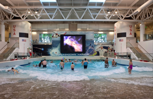 Children and adults enjoy a water play area with waves and a large screen in a bright indoor space.