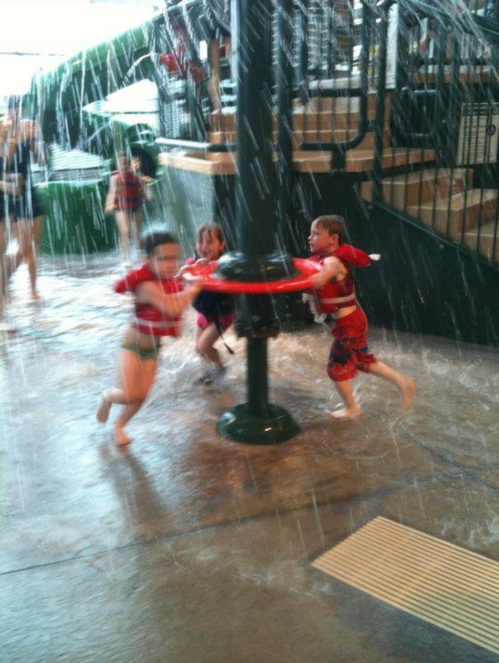 Three children in swimsuits play in a splash area, running around a pole while water sprays down around them.