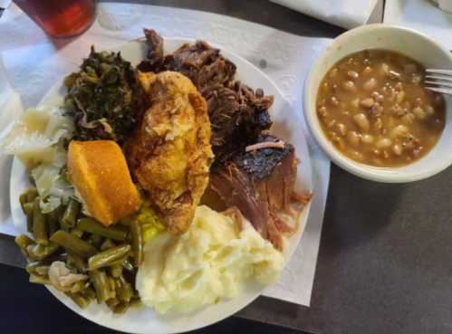 A plate of Southern comfort food featuring fried chicken, pulled pork, greens, cornbread, mashed potatoes, and beans.