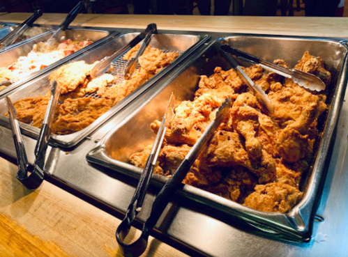 A buffet display featuring trays of crispy fried chicken with tongs for serving.