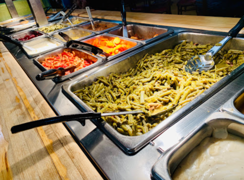 A buffet-style serving station with various dishes, including green beans, carrots, and sauces in metal trays.