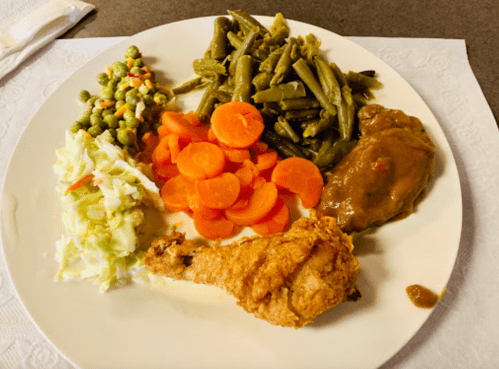 A plate of food featuring fried chicken, green beans, carrots, coleslaw, peas, and brown gravy.