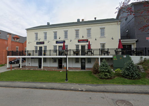 A two-story building with a white exterior, featuring a patio with red umbrellas and a sign for a tavern.