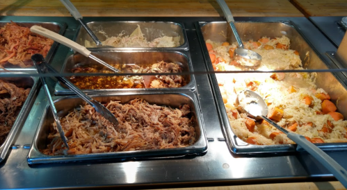 A buffet with several trays of pulled meat, rice, and vegetables, served with metal tongs and spoons.