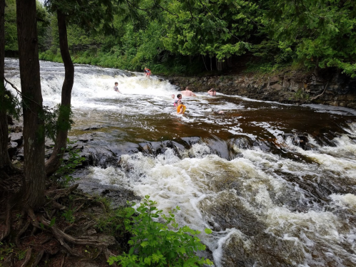 People playing in a river with cascading water, surrounded by lush greenery and rocky banks.