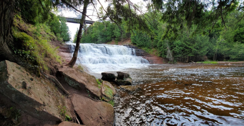 A serene waterfall cascades over rocks, surrounded by lush greenery and a bridge in the background.