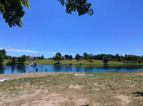 A sunny day at a lake with people swimming and relaxing on the shore, surrounded by green trees and blue sky.