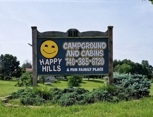 Sign for Happy Hills Campground and Cabins, featuring a smiley face and contact number, set in a green landscape.
