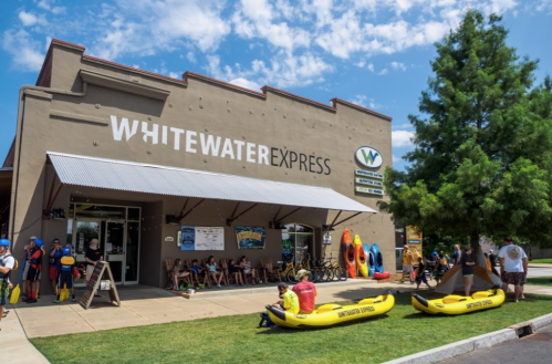 A storefront of Whitewater Express with kayaks outside and people sitting on benches, enjoying a sunny day.