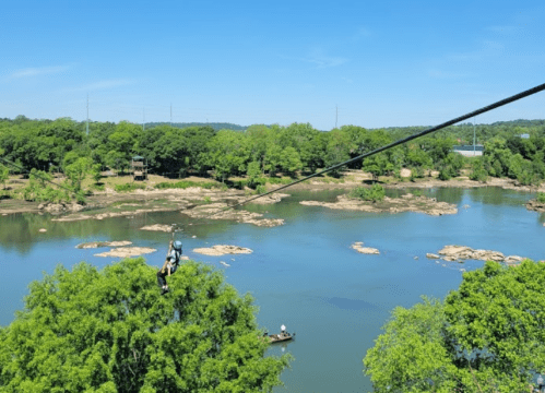 A person zip-lining over a river surrounded by lush greenery and rocky outcrops on a sunny day.