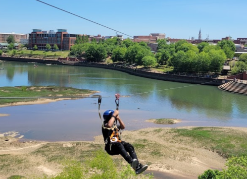 A person zip-lining over a river with a city skyline and green trees in the background on a sunny day.