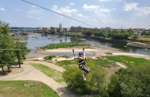 A person zip-lining over a river with greenery and a city skyline in the background on a sunny day.