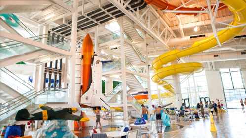 Indoor water park featuring a space shuttle, a yellow water slide, and various attractions with people enjoying the space.
