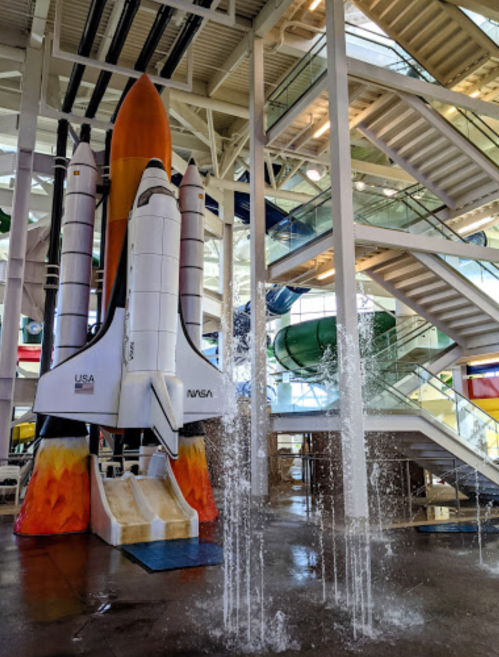 A colorful rocket slide inside a building, with water fountains and stairs in the background.