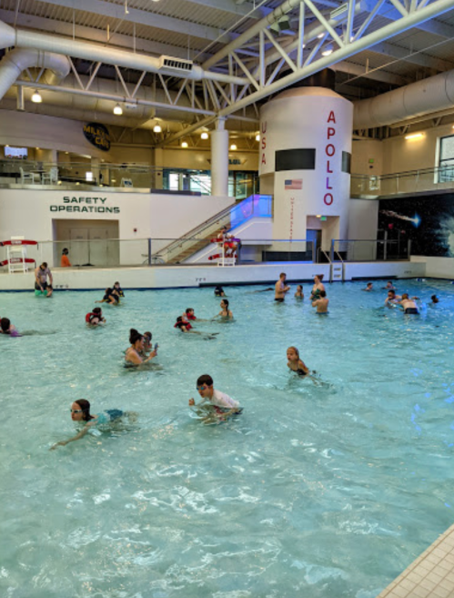 A busy indoor swimming pool with children and adults enjoying the water, surrounded by safety signage.