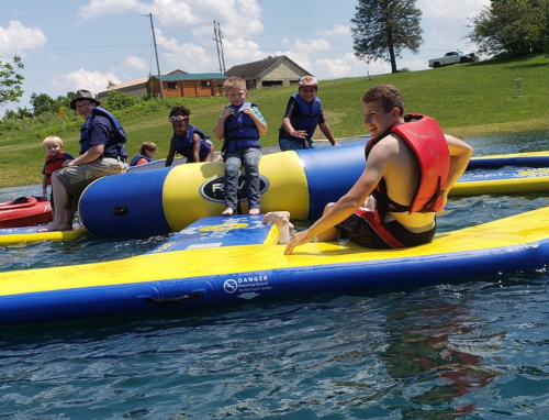 A group of children and adults in life jackets enjoying a water activity on inflatable equipment at a lake.