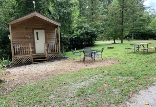 A small wooden cabin with a porch, surrounded by trees, and a picnic table nearby on a grassy area.