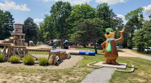 A large cartoon bear statue waves in a park with playground equipment and trees in the background on a sunny day.