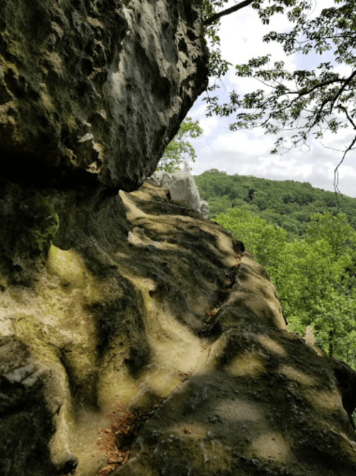 A rocky cliffside with a winding path, surrounded by lush green trees and a cloudy sky in the background.