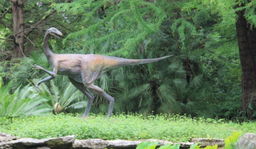 A bronze dinosaur sculpture walking through a lush, green landscape with trees and ferns.