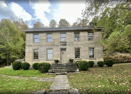 A historic stone house surrounded by greenery and trees, featuring a front staircase and large windows.