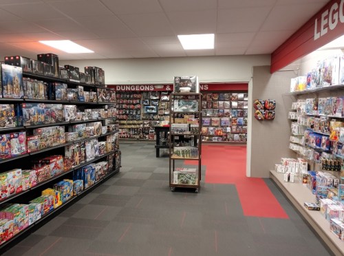 A brightly lit game store aisle filled with shelves of board games, miniatures, and collectibles.
