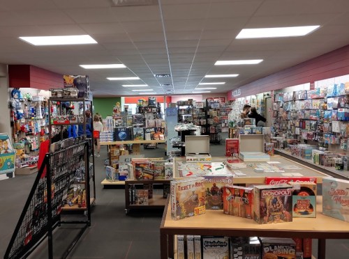 A spacious game store filled with shelves of board games and toys, with customers browsing in the background.