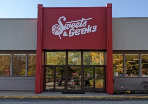 Storefront of "Sweets & Geeks" with large sign, glass doors, and colorful autumn trees in the background.