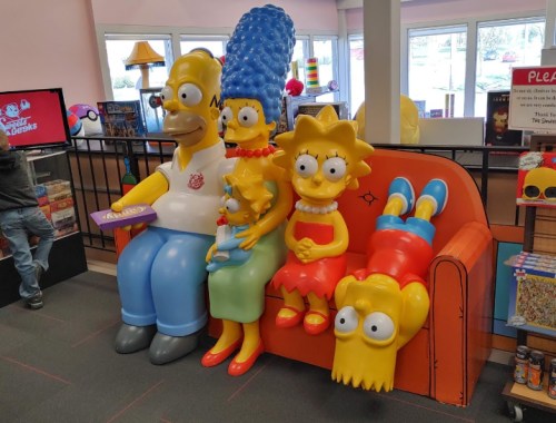 A colorful statue of the Simpson family sitting on a couch in a store, featuring Homer, Marge, Lisa, Maggie, and Bart.