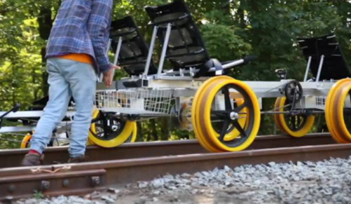 A person adjusts a rail cart with yellow wheels on train tracks surrounded by greenery.