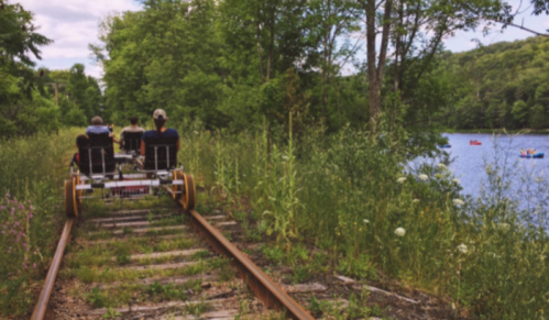 Two people ride a rail bike on overgrown tracks beside a lake, with kayakers visible in the water.