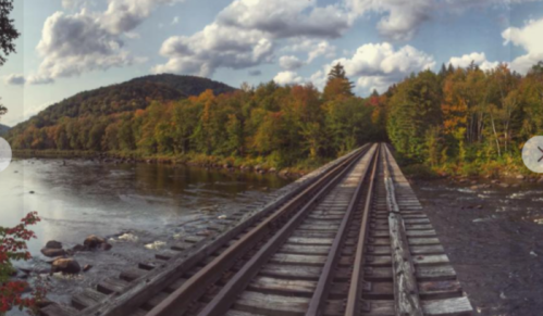 A scenic view of a wooden railway bridge over a river, surrounded by colorful autumn trees and mountains.