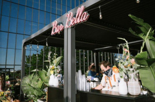 A vibrant outdoor bar with a sign reading "Bar Bella," featuring floral decorations and a bartender in a mask.